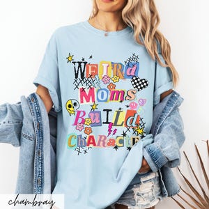 May include: Light blue t-shirt with the text "Weird Moms Build Character" in colorful, playful lettering. The design includes flowers, stars, a checkered heart, and a melting skull. The shirt is a casual, comfortable style.