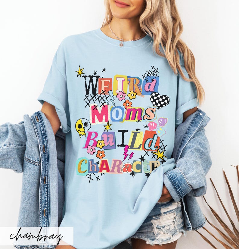 May include: Light blue t-shirt with the text "Weird Moms Build Character" in colorful, playful lettering. The design includes flowers, stars, a checkered heart, and a melting skull. The shirt is a casual, comfortable style.