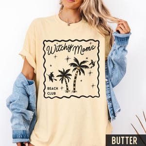 K&ouml;nnte beinhalten: Hellgelbes T-Shirt mit schwarzem Aufdruck &bdquo;Witchy Moms Beach Club&ldquo;. Das Design zeigt Palmen, Sterne und Hexensilhouetten. Das T-Shirt hat kurze &Auml;rmel und eine lockere Passform.