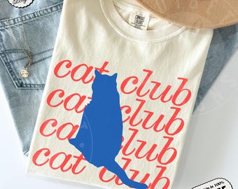 May include: An ivory t-shirt featuring the repeated red text "cat club" in a cursive font. A solid blue silhouette of a sitting cat is centered over the text. The shirt has a crew neckline and a Comfort Colors brand label on the inner collar.
