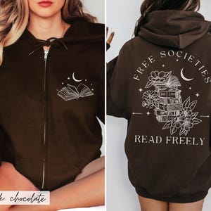 May include: Dark chocolate brown zip-up hoodie with a white graphic of an open book and crescent moon on the front. The back features the text "FREE SOCIETIES" and "READ FREELY" with a stack of books, flowers, and a crescent moon.