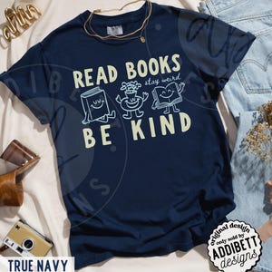 May include: A true navy blue t-shirt featuring a graphic print with the text READ BOOKS stay weird BE KIND. The design includes illustrations of a smiling book, a potted plant, and an open book with arms and legs. The shirt has a crew neckline.