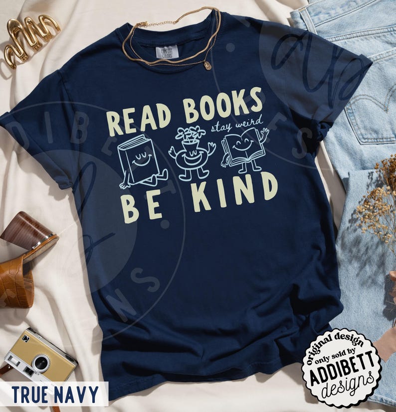 May include: A true navy blue t-shirt featuring a graphic print with the text READ BOOKS stay weird BE KIND. The design includes illustrations of a smiling book, a potted plant, and an open book with arms and legs. The shirt has a crew neckline.