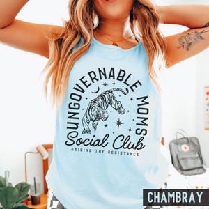 Puede incluir: Camiseta sin mangas azul claro con un gr&aacute;fico de tigre en blanco y negro y el texto "UNGOVERNABLE MOMS Social Club RAISING THE RESISTANCE". La palabra "CHAMBRAY" est&aacute; impresa en la parte inferior.