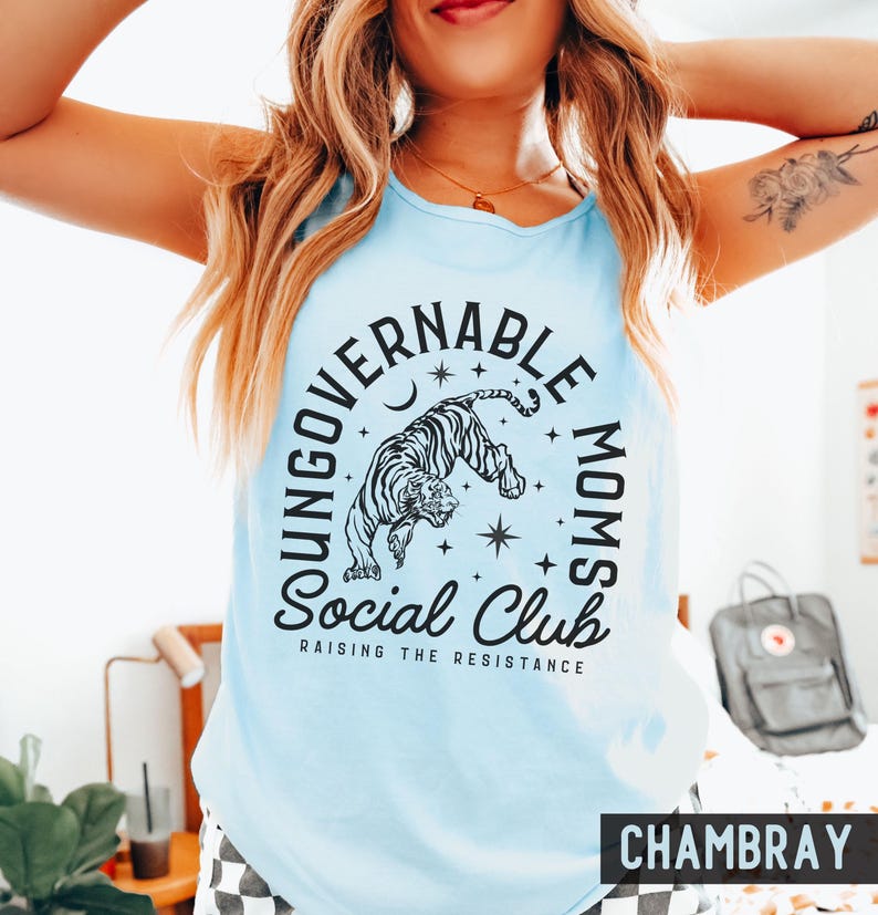 Puede incluir: Camiseta sin mangas azul claro con un gr&aacute;fico de tigre en blanco y negro y el texto "UNGOVERNABLE MOMS Social Club RAISING THE RESISTANCE". La palabra "CHAMBRAY" est&aacute; impresa en la parte inferior.