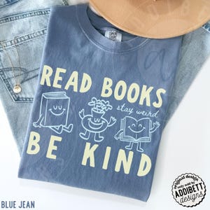 May include: A blue short-sleeve t-shirt with the text READ BOOKS BE KIND in large yellow letters. The design includes a smiling book, a smiling flower vase, and a smiling open book with the script text stay weird in light blue ink.