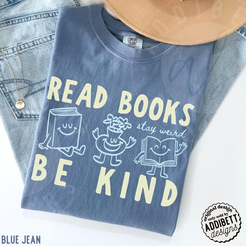May include: A blue short-sleeve t-shirt with the text READ BOOKS BE KIND in large yellow letters. The design includes a smiling book, a smiling flower vase, and a smiling open book with the script text stay weird in light blue ink.