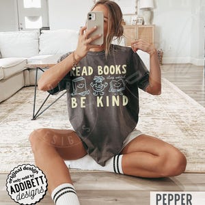 May include: A charcoal gray short-sleeve t-shirt featuring the text READ BOOKS BE KIND in yellow lettering. The design includes three small, light blue cartoon characters: a smiling book, a potted plant, and an open book with a face.