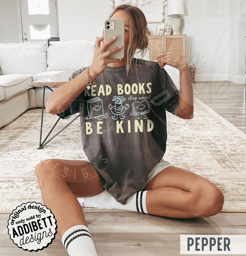 May include: A charcoal gray short-sleeve t-shirt featuring the text READ BOOKS BE KIND in yellow lettering. The design includes three small, light blue cartoon characters: a smiling book, a potted plant, and an open book with a face.