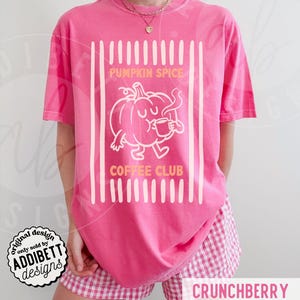 May include: A bright pink short-sleeve t-shirt featuring a white graphic of a cartoon pumpkin holding a coffee mug. The text PUMPKIN SPICE COFFEE CLUB is printed in orange above and below the pumpkin, framed by vertical white stripes.