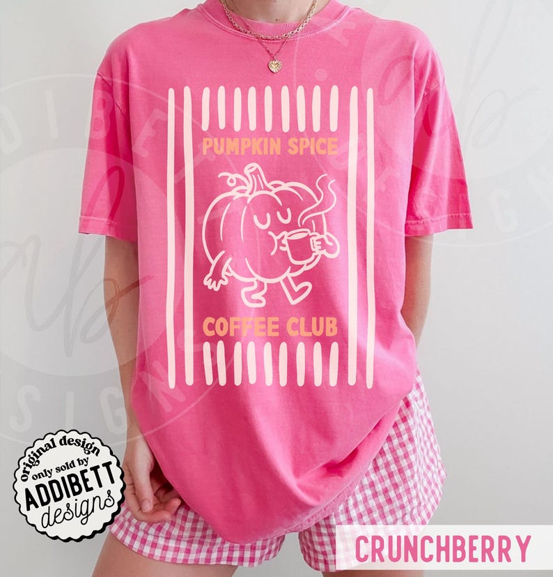 May include: A bright pink short-sleeve t-shirt featuring a white graphic of a cartoon pumpkin holding a coffee mug. The text PUMPKIN SPICE COFFEE CLUB is printed in orange above and below the pumpkin, framed by vertical white stripes.