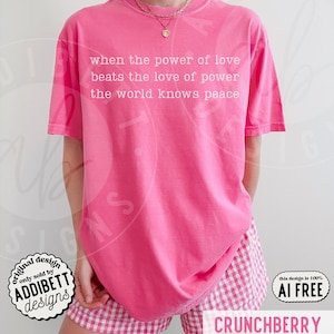 May include: A bright pink short-sleeved t-shirt featuring white text that reads: when the power of love beats the love of power the world knows peace. The shirt is paired with pink and white gingham patterned shorts and a gold chain necklace.