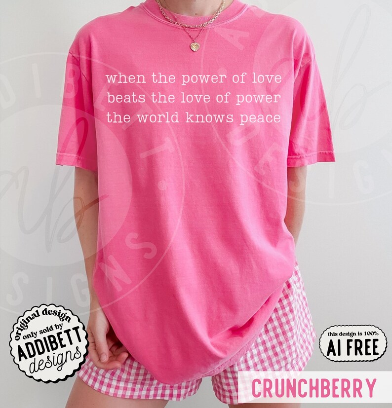 May include: A bright pink short-sleeved t-shirt featuring white text that reads: when the power of love beats the love of power the world knows peace. The shirt is paired with pink and white gingham patterned shorts and a gold chain necklace.