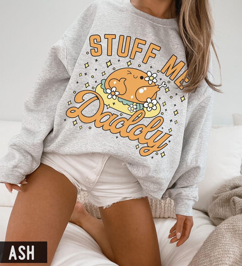 Pu&ograve; includere: Una felpa grigio chiaro con la scritta "STUFF ME Daddy" in lettere arancioni. La grafica presenta un tacchino dei cartoni animati con fiori. La felpa &egrave; uno stile girocollo.