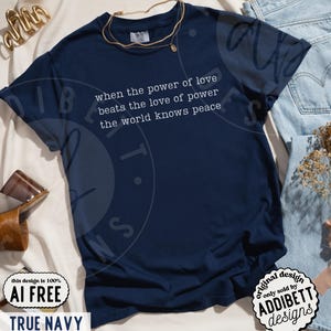 May include: A dark navy blue short-sleeve t-shirt featuring the white printed text: when the power of love beats the love of power the world knows peace. The shirt has a crew neckline and is displayed with a gold-toned necklace and light blue denim.