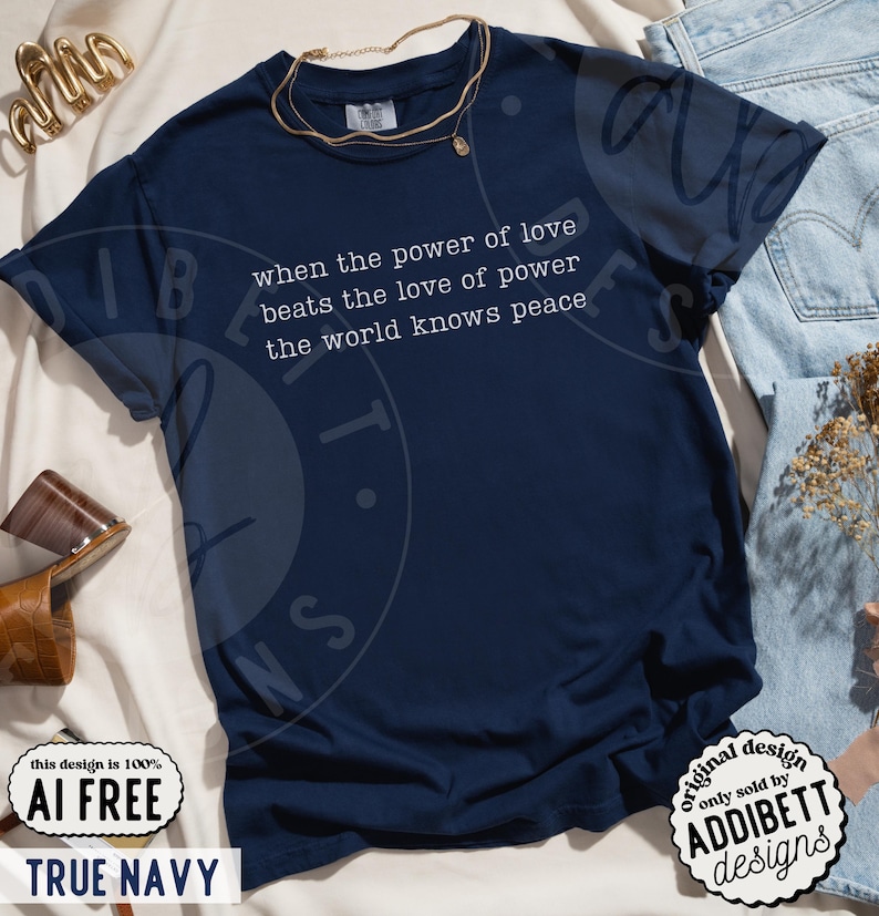 May include: A dark navy blue short-sleeve t-shirt featuring the white printed text: when the power of love beats the love of power the world knows peace. The shirt has a crew neckline and is displayed with a gold-toned necklace and light blue denim.