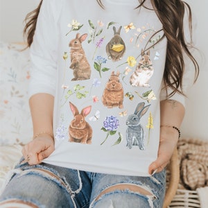May include: White sweatshirt with a colorful floral pattern and multiple brown and gray rabbits. The rabbits are surrounded by flowers, leaves, and butterflies.