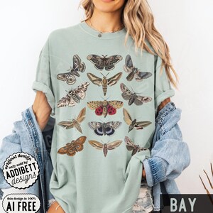 May include: A light sage green t-shirt featuring a graphic print of twelve different moth species arranged in a grid. The moths are depicted in various shades of brown, tan, gray, and cream, with one moth displaying prominent red and black wing patterns.