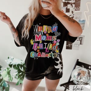 May include: Black t-shirt with the text "Weird Moms Build Character" in colorful, playful lettering. The shirt features floral and lightning bolt graphics. The model is wearing black shorts.
