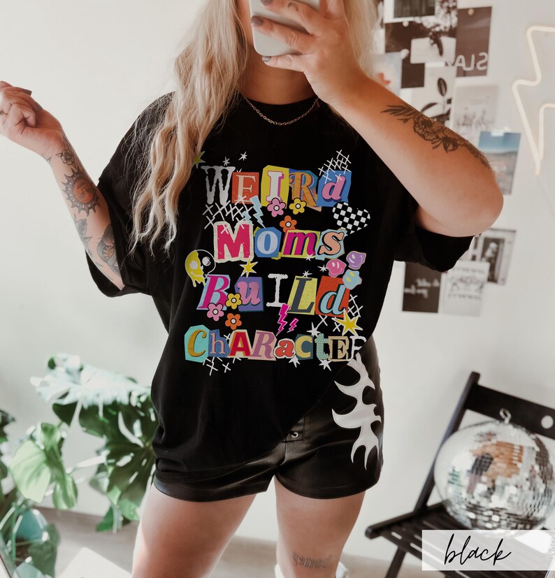 May include: Black t-shirt with the text "Weird Moms Build Character" in colorful, playful lettering. The shirt features floral and lightning bolt graphics. The model is wearing black shorts.