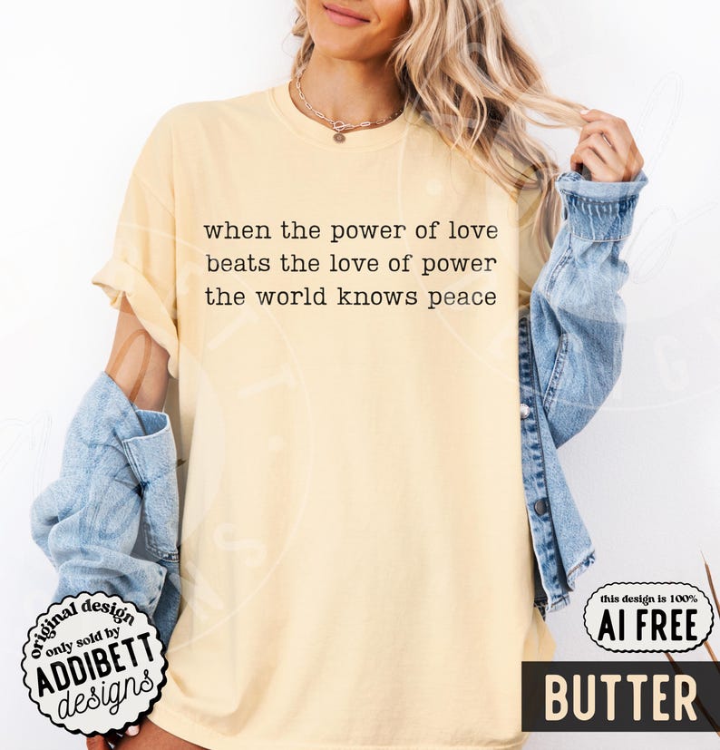 May include: A pale yellow short-sleeve t-shirt featuring black typewriter-style text that reads: when the power of love beats the love of power the world knows peace. The shirt has a crew neckline and is worn with a light blue denim jacket.
