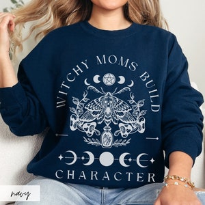 K&ouml;nnte beinhalten: Marineblaues Sweatshirt mit wei&szlig;em Grafikdesign. Das Design umfasst eine Motte, Halbmonde, Sterne und den Text "WITCHY MOMS BUILD CHARACTER". Das Wort "navy" ist unten links aufgedruckt.