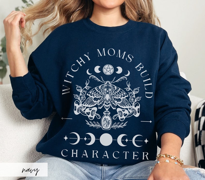 K&ouml;nnte beinhalten: Marineblaues Sweatshirt mit wei&szlig;em Grafikdesign. Das Design umfasst eine Motte, Halbmonde, Sterne und den Text "WITCHY MOMS BUILD CHARACTER". Das Wort "navy" ist unten links aufgedruckt.