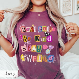 May include: A purple t-shirt with a graphic design that says "Read Books Be Kind Stay Weird" in colorful, cut-out letters.