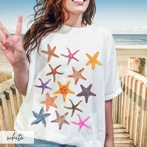 May include: White t-shirt featuring a collection of colorful starfish illustrations. The starfish are in various colors, including orange, pink, purple, and blue. The word "white" is printed at the bottom of the shirt.