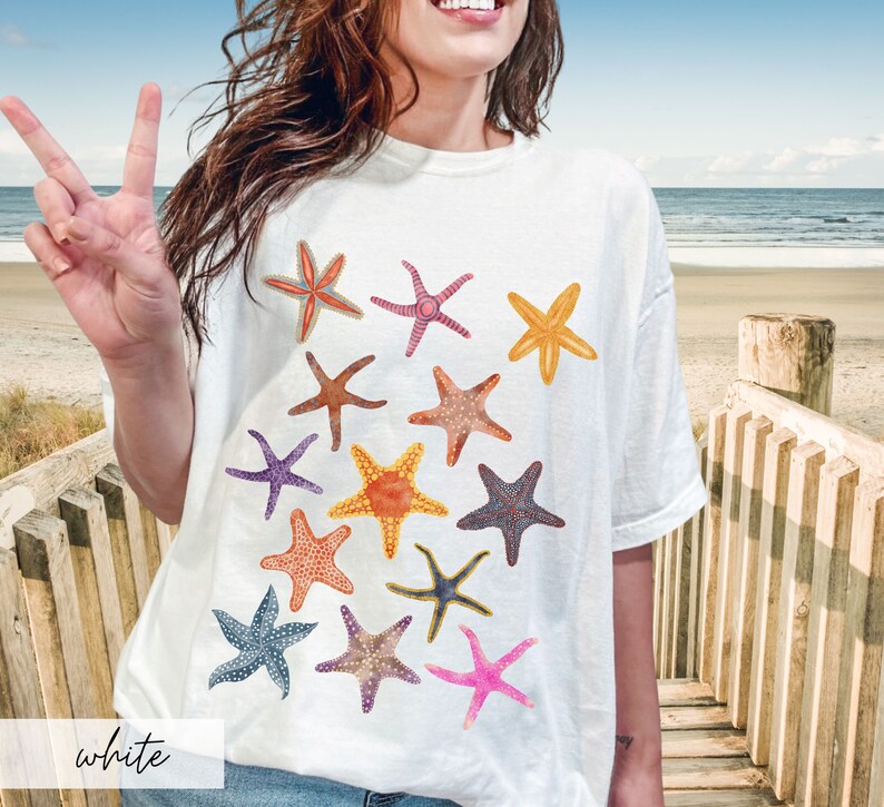 May include: White t-shirt featuring a collection of colorful starfish illustrations. The starfish are in various colors, including orange, pink, purple, and blue. The word "white" is printed at the bottom of the shirt.