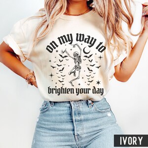 May include: Ivory t-shirt featuring a black graphic of a dancing skeleton, bats, stars, and the text "on my way to brighten your day." The shirt is a casual style, suitable for everyday wear.