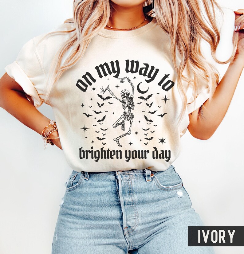 May include: Ivory t-shirt featuring a black graphic of a dancing skeleton, bats, stars, and the text "on my way to brighten your day." The shirt is a casual style, suitable for everyday wear.