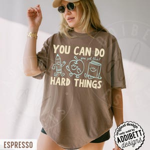 K&ouml;nnte beinhalten: Braunes T-Shirt mit dem Aufdruck "YOU CAN DO HARD THINGS" und Cartoon-Zeichnungen eines Bleistifts, Apfels und Buches. Das Shirt hat eine lockere Passform und stammt aus der Addibett Designs Kollektion.
