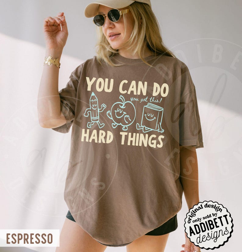 K&ouml;nnte beinhalten: Braunes T-Shirt mit dem Aufdruck "YOU CAN DO HARD THINGS" und Cartoon-Zeichnungen eines Bleistifts, Apfels und Buches. Das Shirt hat eine lockere Passform und stammt aus der Addibett Designs Kollektion.