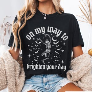 May include: Black t-shirt featuring a dancing skeleton design, bats, stars, and the phrase "on my way to brighten your day." The top is a crew neck style.