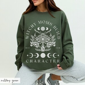 K&ouml;nnte beinhalten: Milit&auml;rgr&uuml;nes Sweatshirt mit dem wei&szlig;en Text "WITCHY MOMS BUILD CHARACTER". Das Design umfasst eine Motte, Mondphasen und botanische Elemente. Das Sweatshirt hat einen Rundhalsausschnitt.