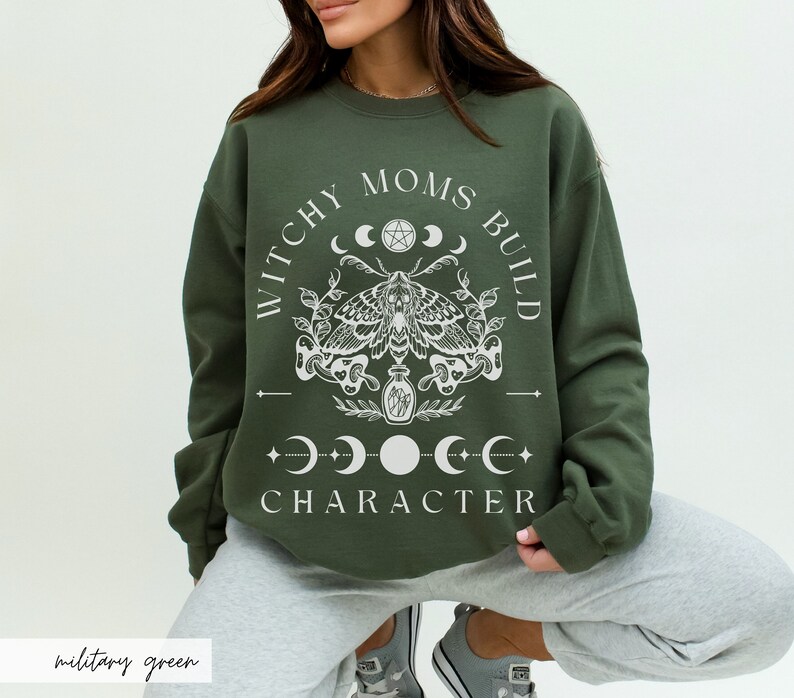 K&ouml;nnte beinhalten: Milit&auml;rgr&uuml;nes Sweatshirt mit dem wei&szlig;en Text "WITCHY MOMS BUILD CHARACTER". Das Design umfasst eine Motte, Mondphasen und botanische Elemente. Das Sweatshirt hat einen Rundhalsausschnitt.