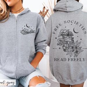 May include: A light gray zip-up hoodie with a black graphic of an open book, moon, and stars on the front. The back features the text "FREE SOCIETIES READ FREELY" with a stack of books, flowers, and a crescent moon.