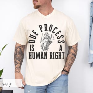 May include: A cream colored t-shirt with a black graphic of a figure holding scales and the text "Due Process is a Human Right".