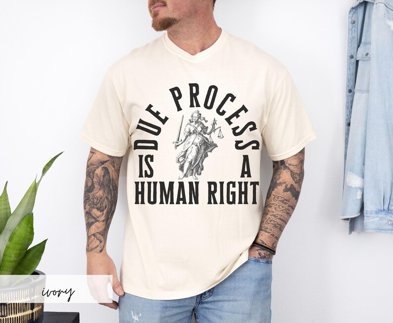 May include: A cream colored t-shirt with a black graphic of a figure holding scales and the text "Due Process is a Human Right".