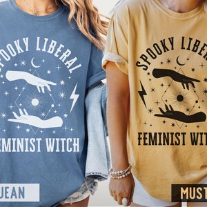 Puede incluir: Dos camisetas, una azul vaquero y otra mostaza, con el texto "Spooky Liberal Feminist Witch". El dise&ntilde;o incluye manos, rayos, estrellas y una luna creciente. Las camisetas son ropa casual.