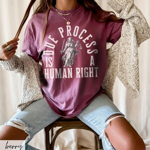 May include: A woman wearing a maroon t-shirt with the text "Due Process is a Human Right" and a graphic of a woman holding scales. She is wearing light blue jeans and a beige cardigan.