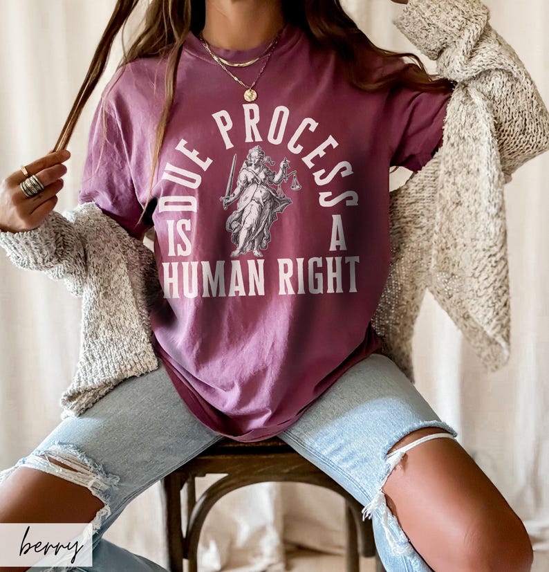 May include: A woman wearing a maroon t-shirt with the text "Due Process is a Human Right" and a graphic of a woman holding scales. She is wearing light blue jeans and a beige cardigan.