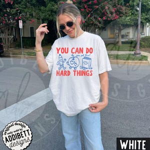 K&ouml;nnte beinhalten: Wei&szlig;es T-Shirt mit dem Text "YOU CAN DO HARD THINGS" in Rot und Blau. Das Design zeigt Cartoon-Illustrationen von Bleistift, Apfel und Buch. Das Shirt hat eine lockere Passform.