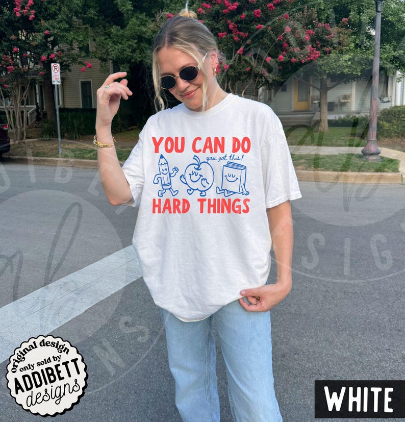 K&ouml;nnte beinhalten: Wei&szlig;es T-Shirt mit dem Text "YOU CAN DO HARD THINGS" in Rot und Blau. Das Design zeigt Cartoon-Illustrationen von Bleistift, Apfel und Buch. Das Shirt hat eine lockere Passform.