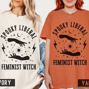 Puede incluir: Camisetas de color marfil y &ntilde;ame con el texto "Spooky Liberal Feminist Witch" en negro. El dise&ntilde;o incluye manos, una luna creciente, estrellas y rayos. Las camisetas son de manga corta y tienen un corte holgado.