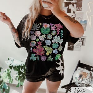 Puede incluir: Camiseta negra con estampado de suculentas de colores. La camiseta tiene un corte holgado y cuello redondo.