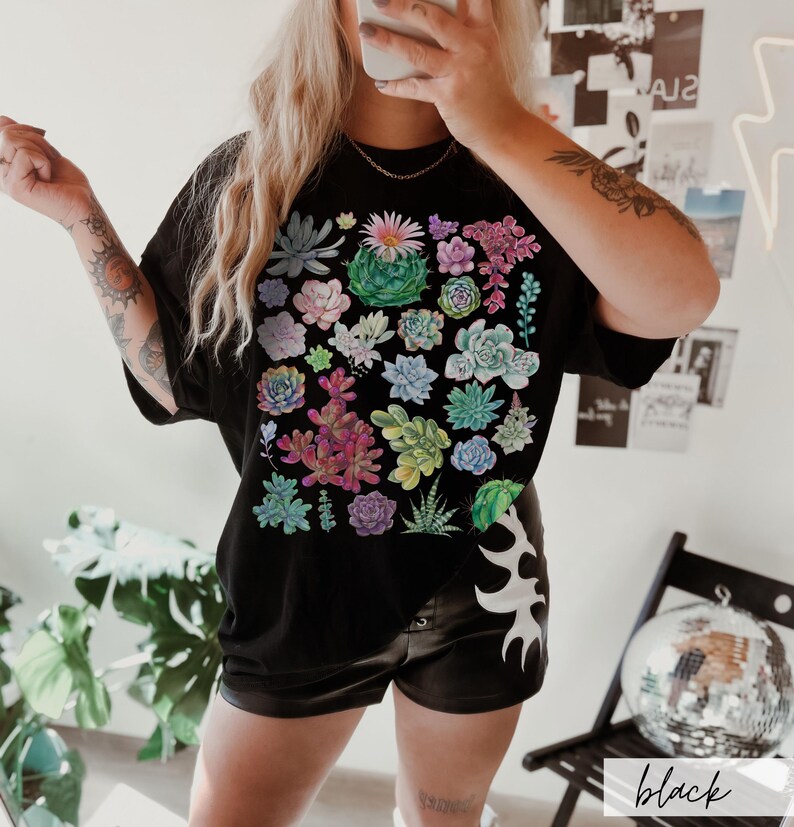 Puede incluir: Camiseta negra con estampado de suculentas de colores. La camiseta tiene un corte holgado y cuello redondo.
