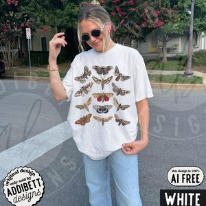 May include: An off-white short-sleeve t-shirt featuring a graphic print of various moth species in shades of brown, tan, and grey. The moths are arranged in a grid-like pattern across the front of the shirt, showcasing diverse wing shapes and intricate patterns.