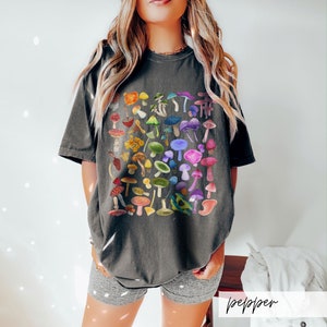 K&ouml;nnte beinhalten: Ein dunkelgraues T-Shirt mit einem bunten Pilzdruck. Die Pilze sind in einem quadratischen Muster angeordnet und in verschiedenen Schattierungen von Rot, Orange, Gelb, Gr&uuml;n, Blau, Lila und Rosa.